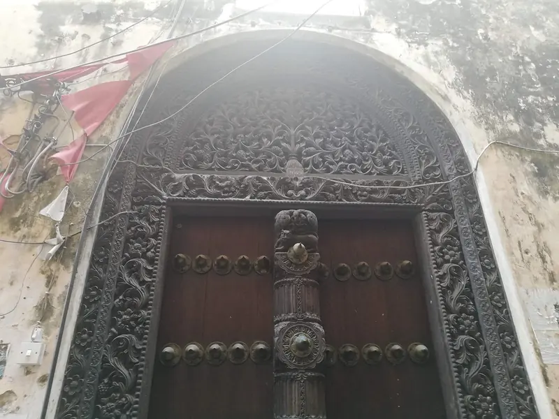 A carved Zanzibar door frames a Stone Town entrance, the studded brass fittings and floral relief typical of the island's Swahili and Indian merchant houses.