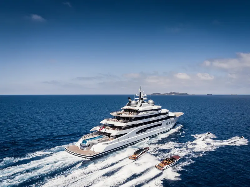 Amadea underway off an island coastline, with two tenders and jet skis in her wake. The 106-metre Lurssen was delivered in 2017 and has since become the highest-profile yacht ever sold at a US government forfeiture auction.