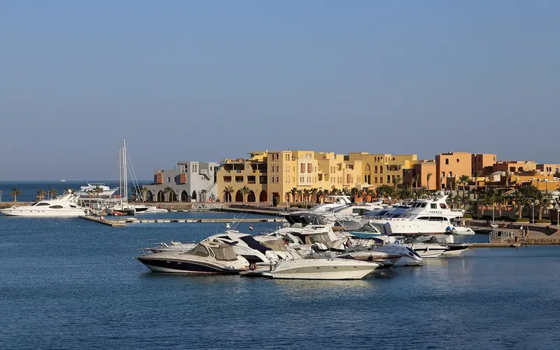 Motor yachts and a sailing yacht on the pontoons in front of Abu Tig's painted waterfront blocks, the sea open behind the breakwater.