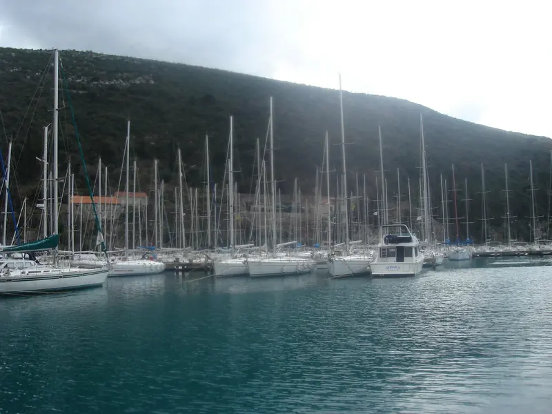 Sailing yachts moored bow-to at ACI Marina Dubrovnik's pontoons in Komolac, with the wooded slope of the Rijeka Dubrovacka inlet behind them.