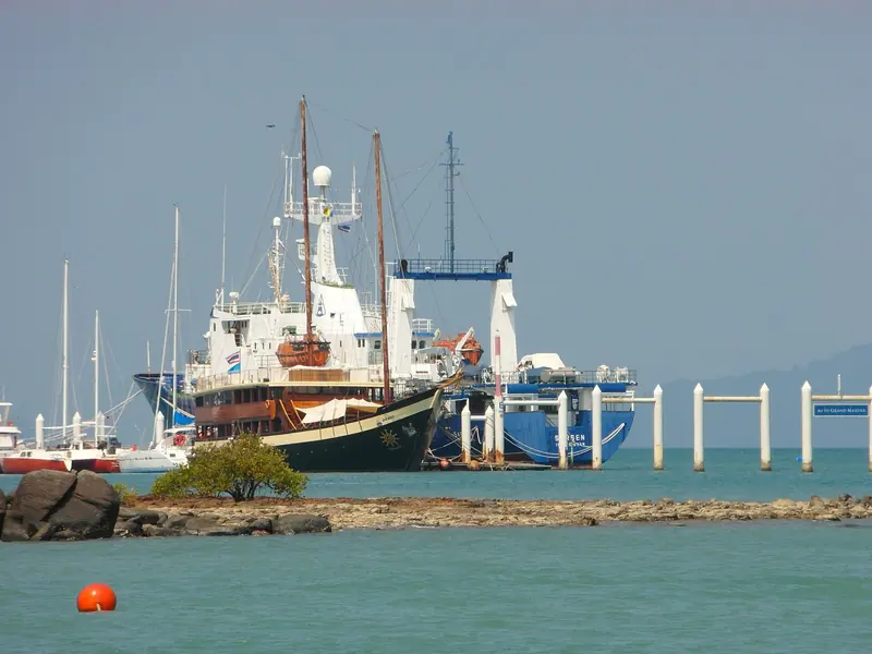 A traditional Thai-style motor yacht and a support vessel moored at Ao Po Grand Marina's outer pontoons, the marina's own sign visible on the piling at right and the hills of Phang Nga Bay behind.