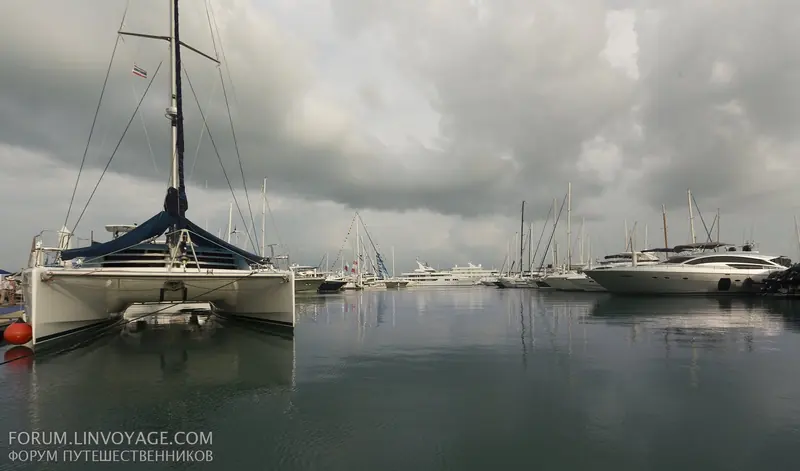 A catamaran's bow in the foreground with motor yachts and sailing yachts lined along Ao Po Grand Marina's basin under a northeast-monsoon sky.