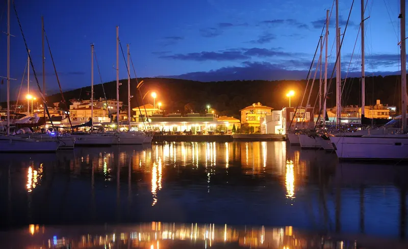 Cesme Marina at dusk, its basin lit and lined with cruising yachts on the Aegean coast west of Izmir.