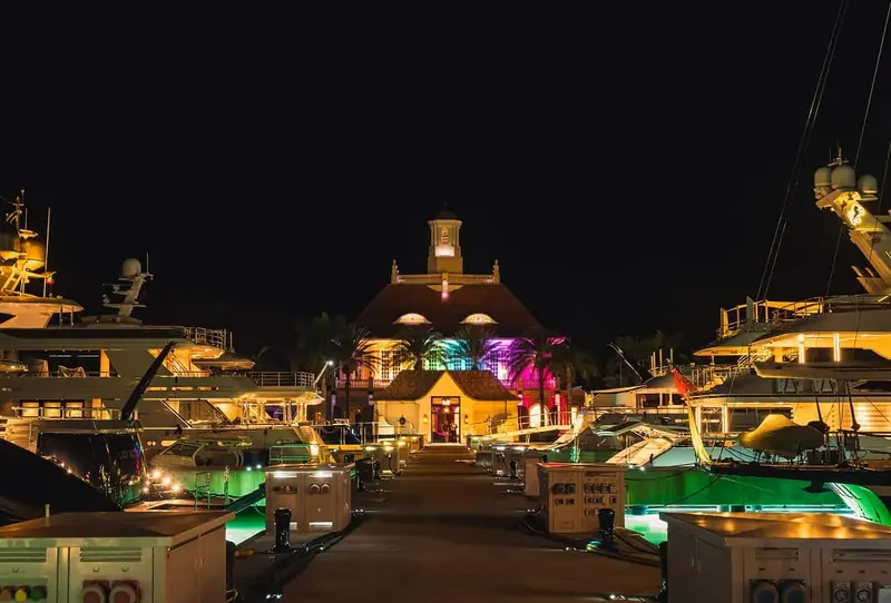 The Marina Village clubhouse lit up at night, seen down the main dock between two berthed superyachts.