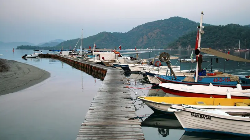 A wooden pontoon reaching into the still water of Gocek bay at first light, local fishing boats made fast alongside.