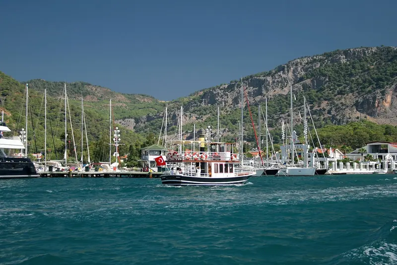 A day-tripper boat crosses the anchorage in front of the marina, gulet masts and the bare limestone ridge above the bay.