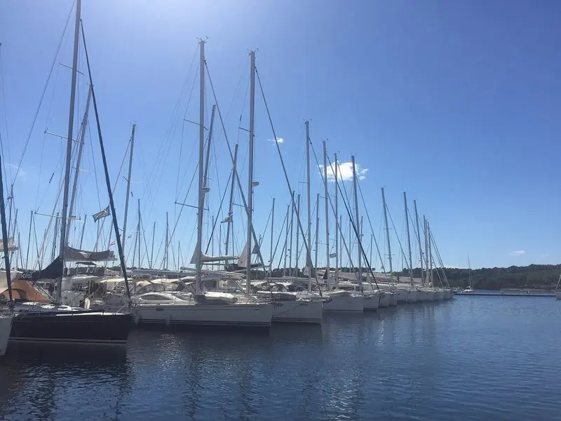 Sailing yachts sit bows-to along one pontoon in flat midday water, the low wooded far shore of the channel closing the view.