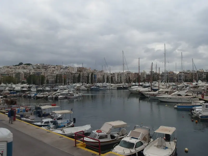 The main basin under a grey Attic sky: sailing yachts, motor cruisers and small open boats share the same pontoons, with the apartment blocks of Kastella rising straight from the waterline behind them.