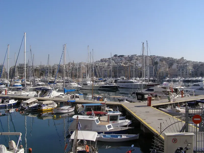 On a clear day the same basin looks like a different marina. Piraeus, Greece's largest passenger port and roughly the third busiest in the Mediterranean, sits close enough that the two skylines merge into one.