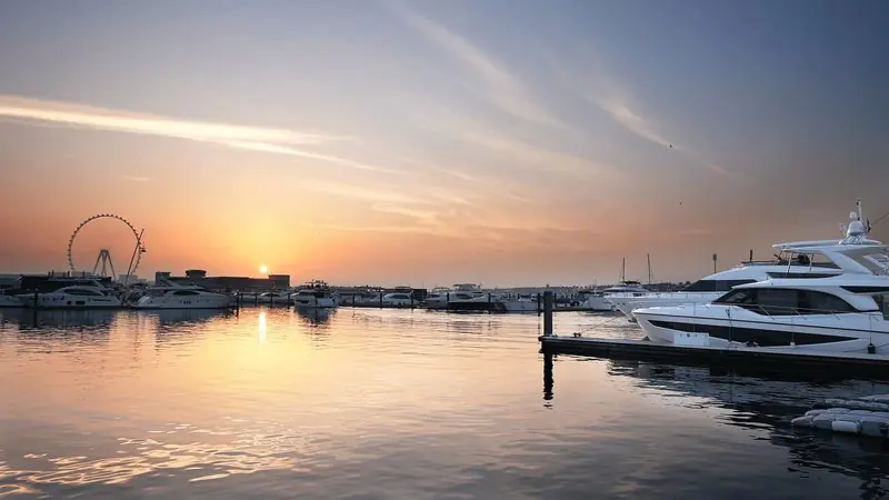 Motor yachts lie along the pontoons while the sun drops behind a giant observation wheel.
