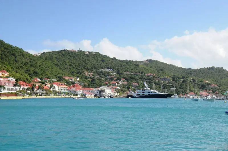 A large motor yacht lies alongside the quay at Gustavia, with the red roofs of the town and the hillside villas rising directly behind her.