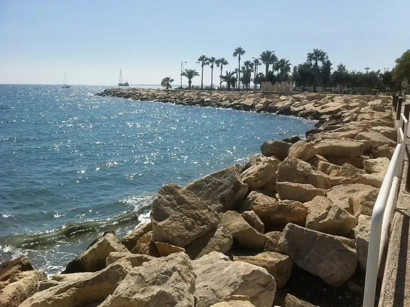 Rock armour walls the seaward edge of the development, with a yacht standing offshore in the open water beyond the palms of the promenade.