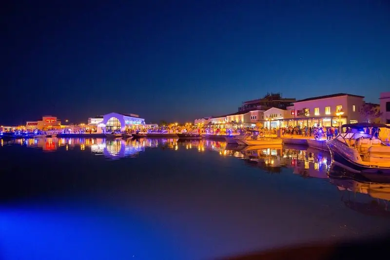 Looking back across the basin at blue hour, the lit arcades and the boats on the near pontoon show a marina built to be walked as much as berthed in.