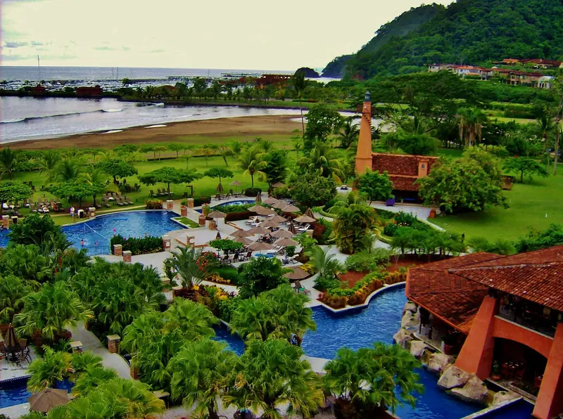 Herradura Bay and the Los Suenos marina basin seen from the resort grounds, with the resort's orange lighthouse-tower landmark and the boats moored inside the breakwater visible across the beach