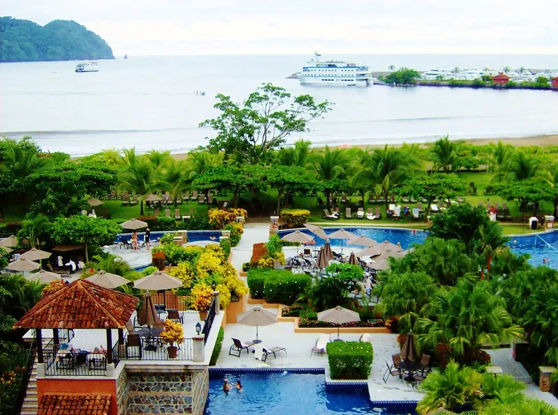 A tour vessel anchored off Herradura Bay with the Los Suenos marina breakwater and moored yachts visible on the point beyond the resort pools and gardens