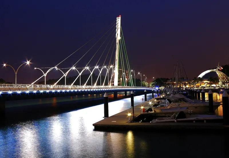 The Keppel Bay Bridge lit up after dark, its cables reflected in the water, with the glass-domed clubhouse and berthed yachts to the right.