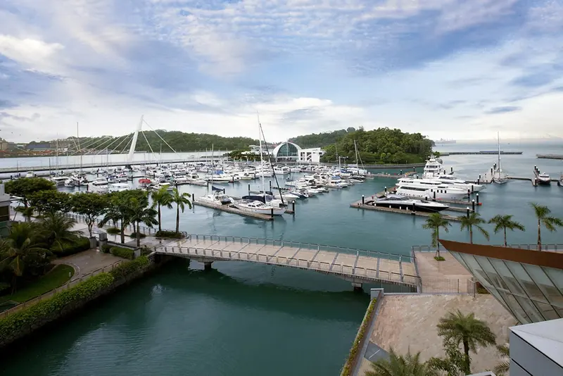 A small footbridge over the tidal canal in front of the marina basin, with berths, the glass-domed clubhouse and superyachts on the far pontoon.