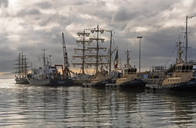 Tall ships moored at sunrise in Doca de Alcantara, with a harbour crane and the 25 de Abril bridge in the background.