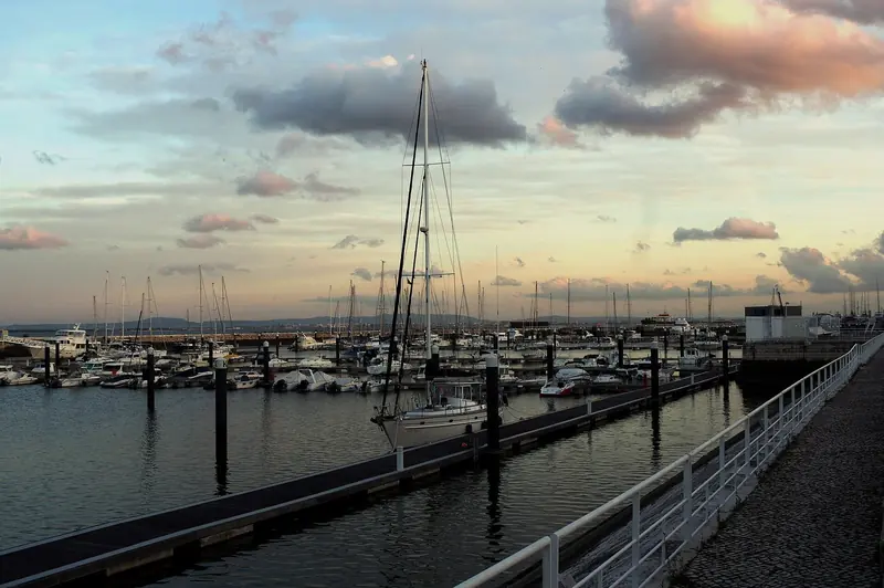 A yacht ties up alone on an empty finger pontoon as the sky over the marina turns pink at dusk.