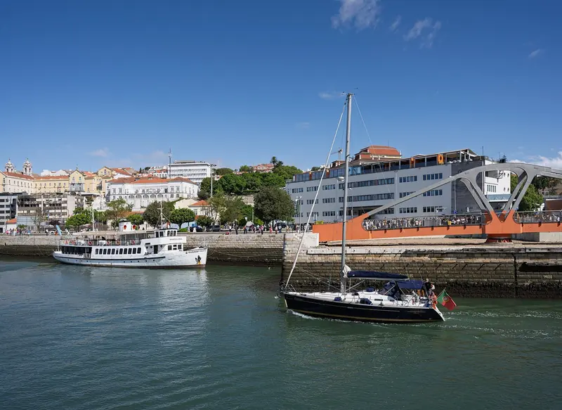 The Beneteau Oceanis 393 Pedras I enters Doca de Alcantara under sail, passing the dock's arched pedestrian bridge.