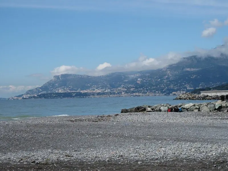 West of the beach the coast runs on toward Cap Martin, with the high buildings of Monaco visible on the skyline.