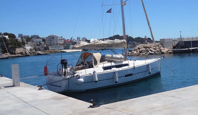 A charter sloop lies bow out on the Calanova quay, with the harbour mouth and its red beacon just beyond the breakwater rocks.