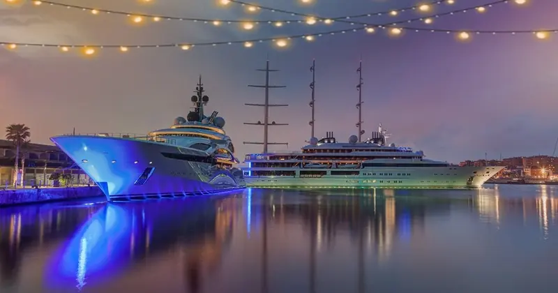Yachts lit at dusk on flat water, seen from the quayside under strings of festoon lights.