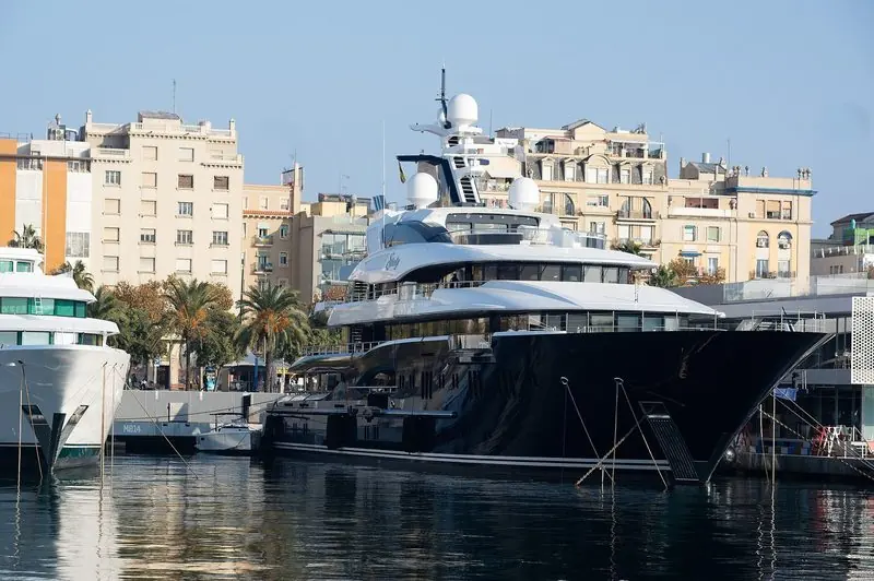 A dark-hulled motor yacht sits alongside the main superyacht berth, with the palm-lined promenade and the marina buildings a minute from the passerelle.
