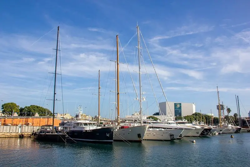Classic motor and sailing yachts moor stern-to the old harbour wall, with the aquarium and the Maremagnum buildings a short walk behind them.