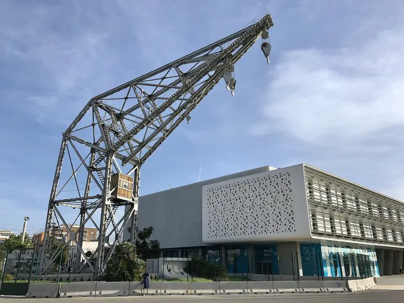 A preserved dockside gantry crane from the historic port stands beside the modern Oceanografic-district building on the marina's northern edge, a reminder of the working harbor the America's Cup basin was built inside.