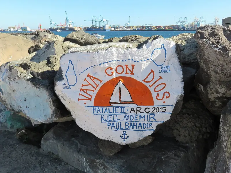 A crew's hand-painted rock for Natalie II, ARC 2015, tracing the route from Gran Canaria to St Lucia, with the container terminal cranes of the Port of Las Palmas behind.