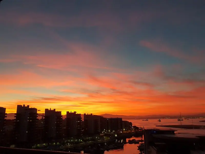 Sunset over the apartment towers, with the marina basin and its boats tucked in below and the Spanish shore closing the horizon.