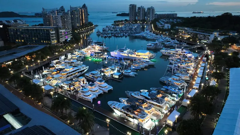 The marina at dusk, the pontoons and the boats lit, the strait and its anchored shipping beyond the entrance.