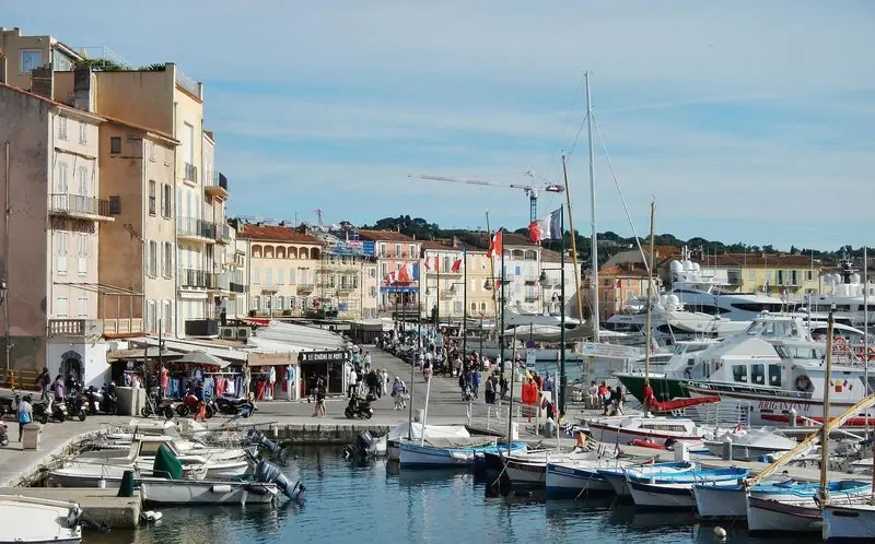 Crowds move along the quay past the shopfronts and awnings while tenders and workboats fill the near pontoons, the harbour at its summer busiest.