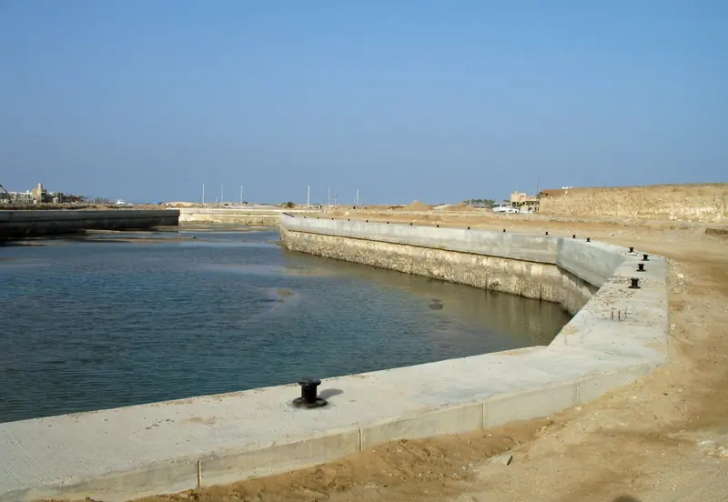 A new basin under construction at Port Ghalib in January 2023, its concrete quay wall and mooring bollards already in, existing yacht masts visible in the marina beyond.