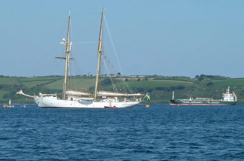A two masted schooner rides at anchor in the roads with a coaster lying further out and the wooded shore beyond.