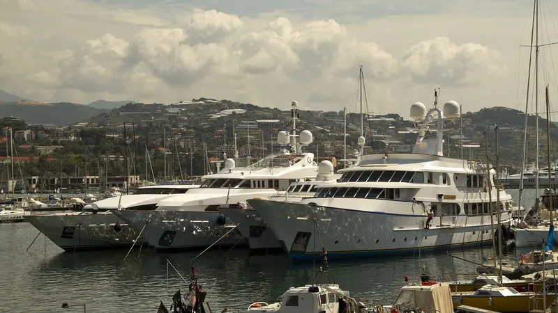 Megayachts on the quay at Portosole, radar domes and satellite antennas above the superstructure, the Ligurian hills behind.