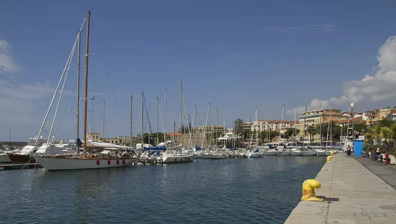 Classic and modern sailing yachts moored along a Portosole quay, the marina promenade and the town behind.