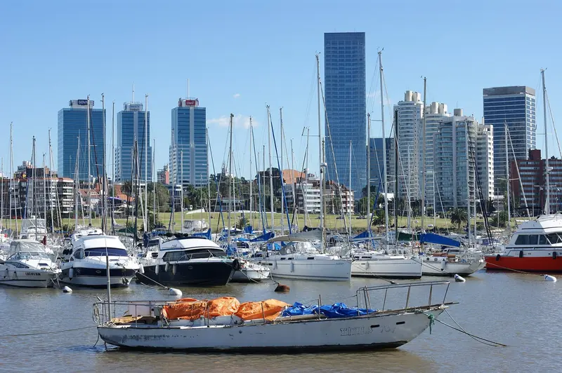 Ground level among the berths: cruisers and sailboats crowd the inner basin ahead of Montevideo's Bulevar Artigas office towers, with a working wooden fishing boat riding at anchor in the foreground.