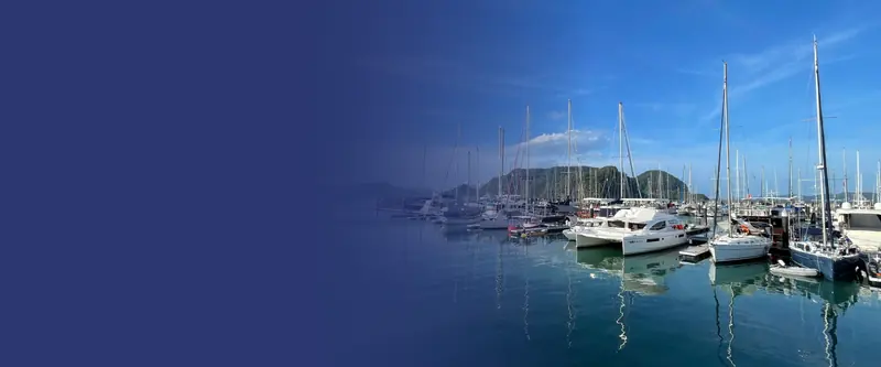 A catamaran and cruising sailboats moored on a finger pontoon at dusk, the Langkawi hills across the anchorage.
