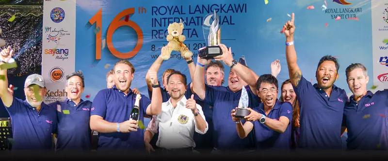 A winning crew lifts the trophy on stage at the 16th Royal Langkawi International Regatta, the club's own banner behind them.