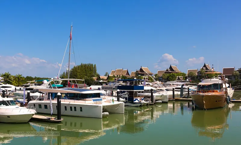 Motor yachts and a catamaran on the pontoons in daylight, the marina's Thai-pavilion clubhouse roofline behind, a very different setting from Ao Po's open bay further north.