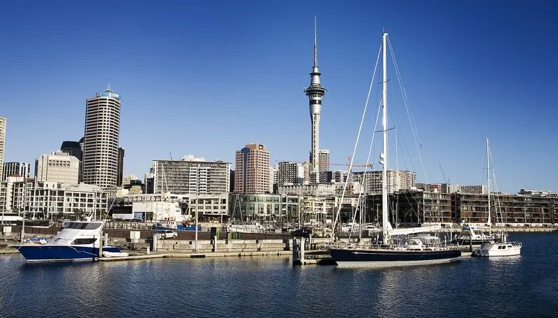A large blue-hulled sloop lies on the outer pontoon of the Wynyard basin, the city towers and the Sky Tower standing directly behind the berths.