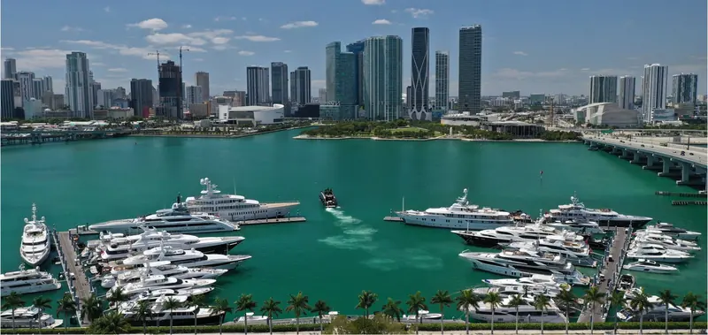 A yacht heads out past the marina's palm-lined pontoons, downtown Miami's towers and construction cranes across the water.
