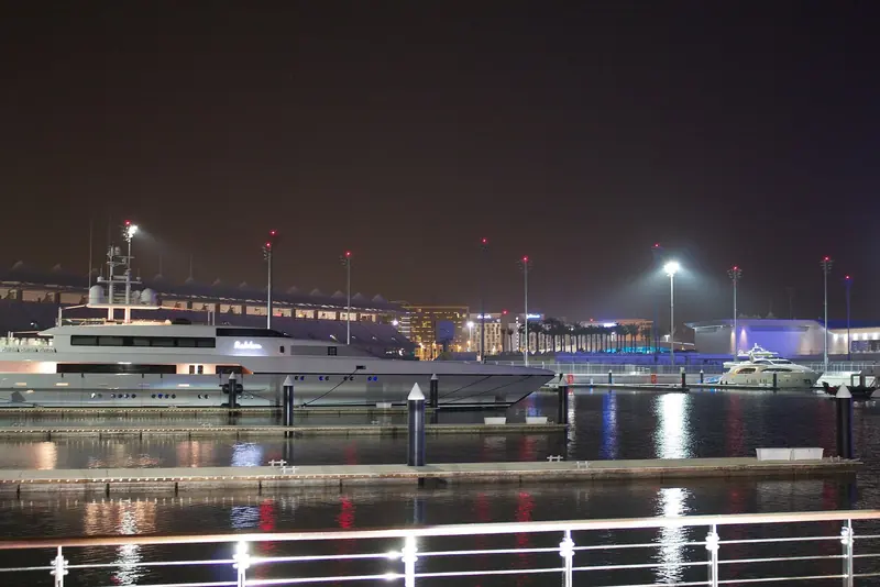 A large yacht lies on the marina berths after dark, the tented roof of the grandstand and its floodlights standing behind her.