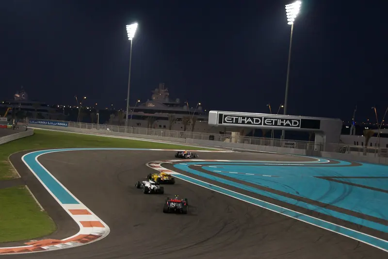 Race night on the circuit, cars running through the turn under the floodlights with a yacht berthed just the other side of the barrier.