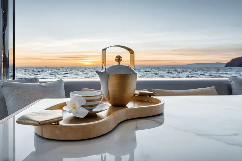 A gold-finished tea service laid on the cockpit table at sunset, the hills of the Gulf of Genoa in silhouette.