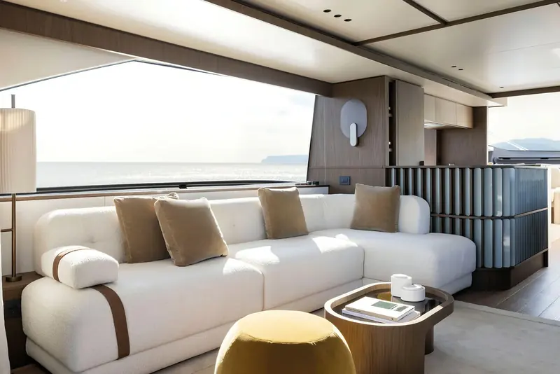 The main-deck saloon at last light, low boucle seating catching the sun off the water and a slatted timber-clad galley counter dividing it from the day area.