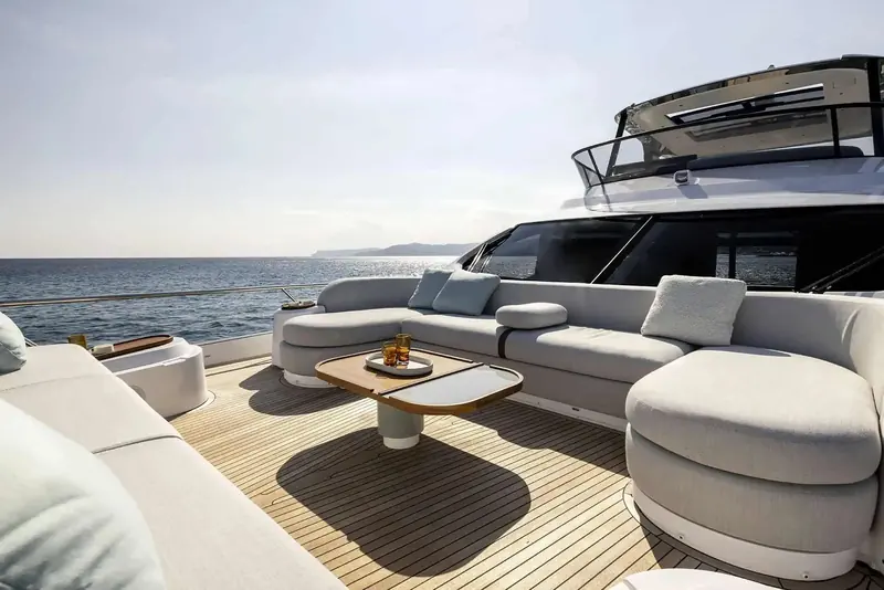The flybridge lounge in full sun, drinks left on a floating tray table and a coastline visible off the port rail.
