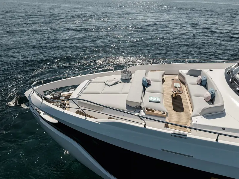 The foredeck lounge underway, sun catching the wake astern and a folded chart on the sofa - the kind of open-water forward seating that a full-beam owner's suite below makes possible.